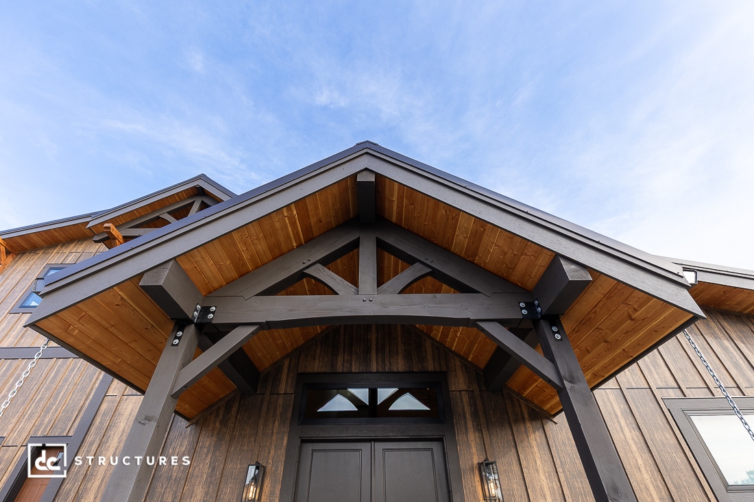 A low-angle view of a modern wooden building entrance with a dark timber gabled porch, natural wood paneling, large double doors, and a blue sky overhead.