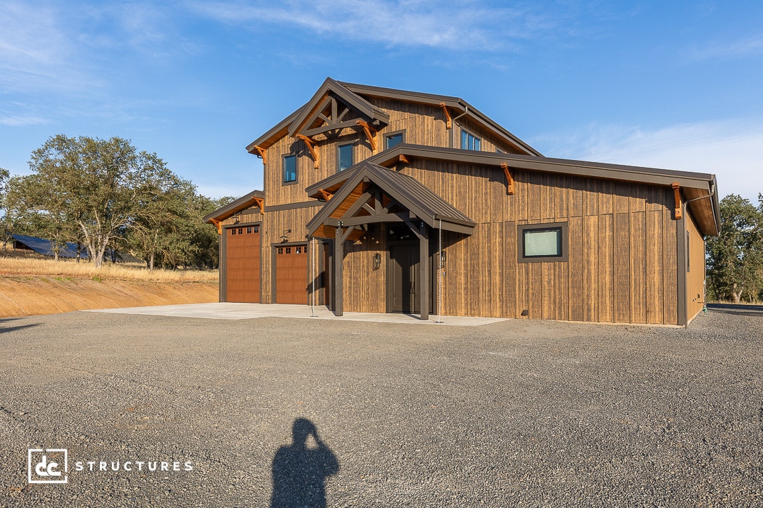 A modern two-story wooden building with large garage doors, black trim, and exposed beams sits on a gravel lot under a clear blue sky. Trees and dry grass line the background. A shadow of a person is visible in the foreground.