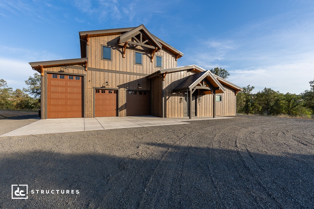 A modern two-story wood-paneled house with two large garage doors sits on a gravel lot, surrounded by trees under a blue sky. The house features rustic architectural details and a concrete driveway.