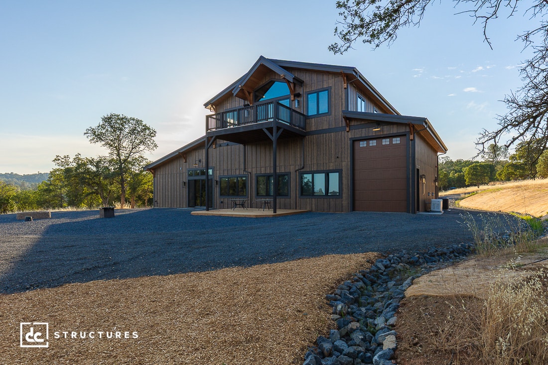 A modern two-story barn-style house with wood siding, large windows, a balcony, and a garage door, set on a gravel lot surrounded by trees and hills under a clear sky.