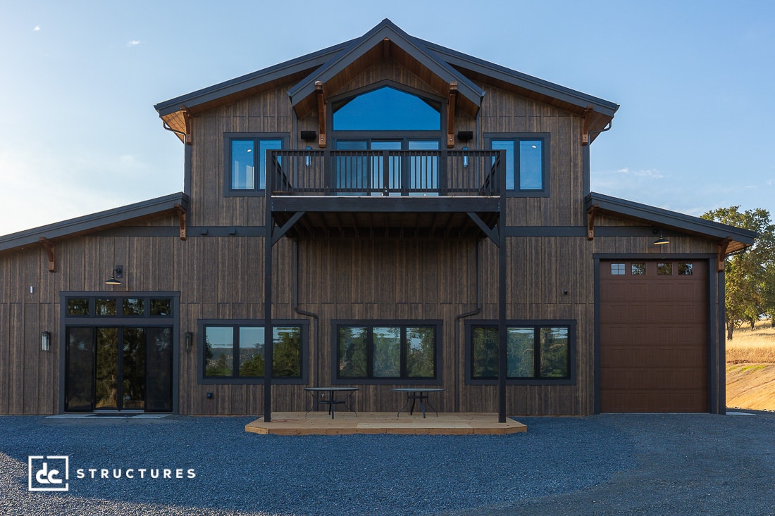 A modern barn-style building with dark wood siding, large windows, a glass balcony, and a tall garage door. The front features a small patio area with a bench, and the ground is covered with gravel.