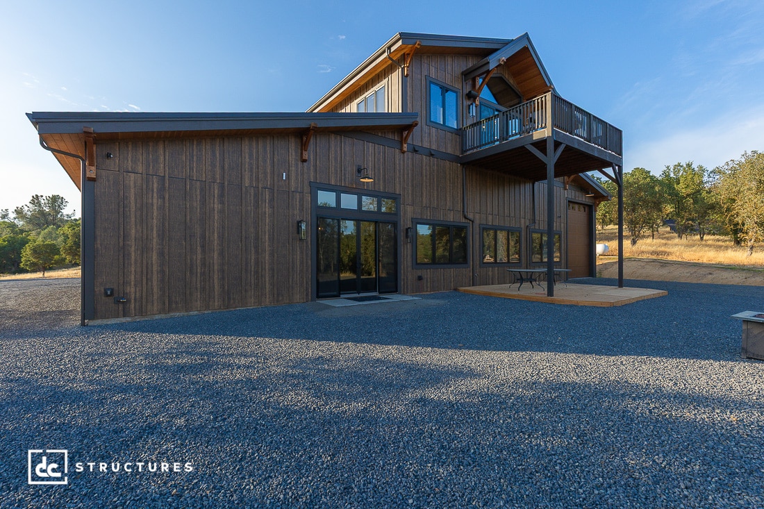 A modern two-story building with wood siding, large windows, and an upper balcony pictured on a gravel lot with trees and grass in the background. The sky is clear and sunny.