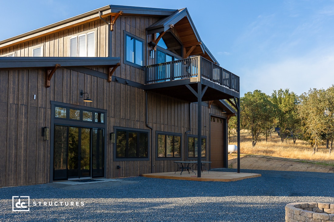 A modern two-story barn-style building with large windows, a wooden exterior, a balcony, and a small patio area, set on gravel with trees and dry grass in the background under a blue sky.