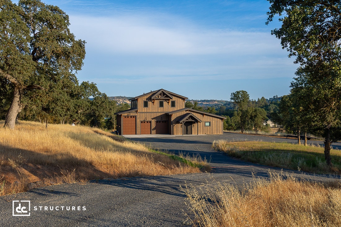 A modern barn-style house with a large garage sits at the end of a curving driveway, surrounded by tall trees and dry golden grass under a clear blue sky.
