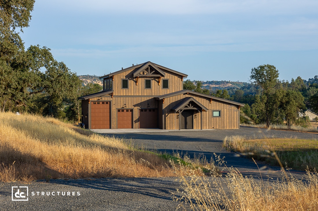 A large, modern barn-style house with wooden siding, three garage doors, and a gravel driveway sits in a rural landscape surrounded by trees and dry grass under a clear sky.