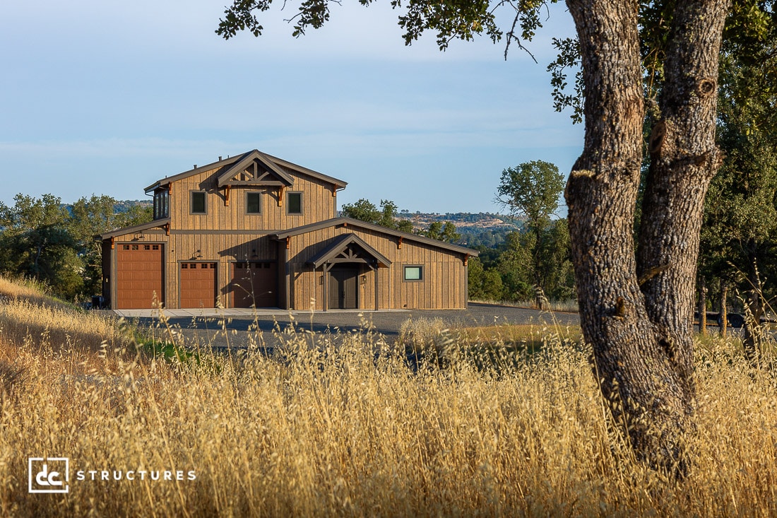 A modern barn-style house with wooden exterior and large garage doors sits surrounded by dry golden grass and trees under a clear sky.