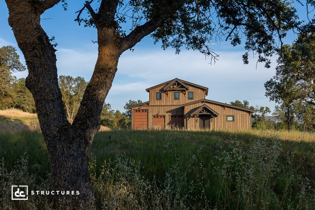 A rustic two-story wooden house stands in a grassy, tree-dotted landscape under a blue sky, partially framed by a large tree in the foreground.