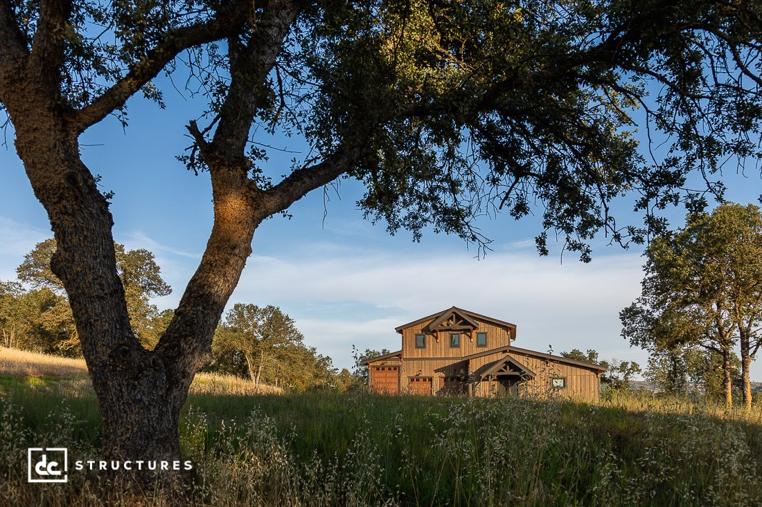 A rustic wooden barn-style house stands in a grassy field surrounded by trees, with sunlight casting warm tones across the landscape. A large tree frames the left side of the image.
