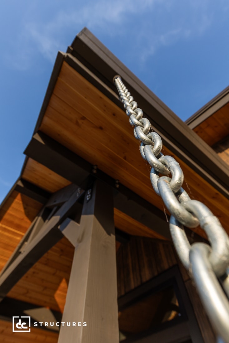 Close-up view looking up at a metal rain chain attached to the roof of a wooden building, with blue sky in the background.