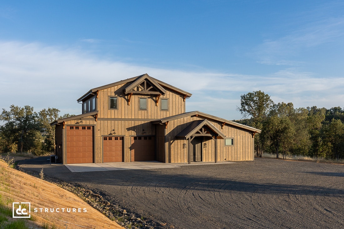 Two-story barn-style house with wood siding, three garage doors, and an arched porch, set on a gravel lot surrounded by trees under a clear blue sky.