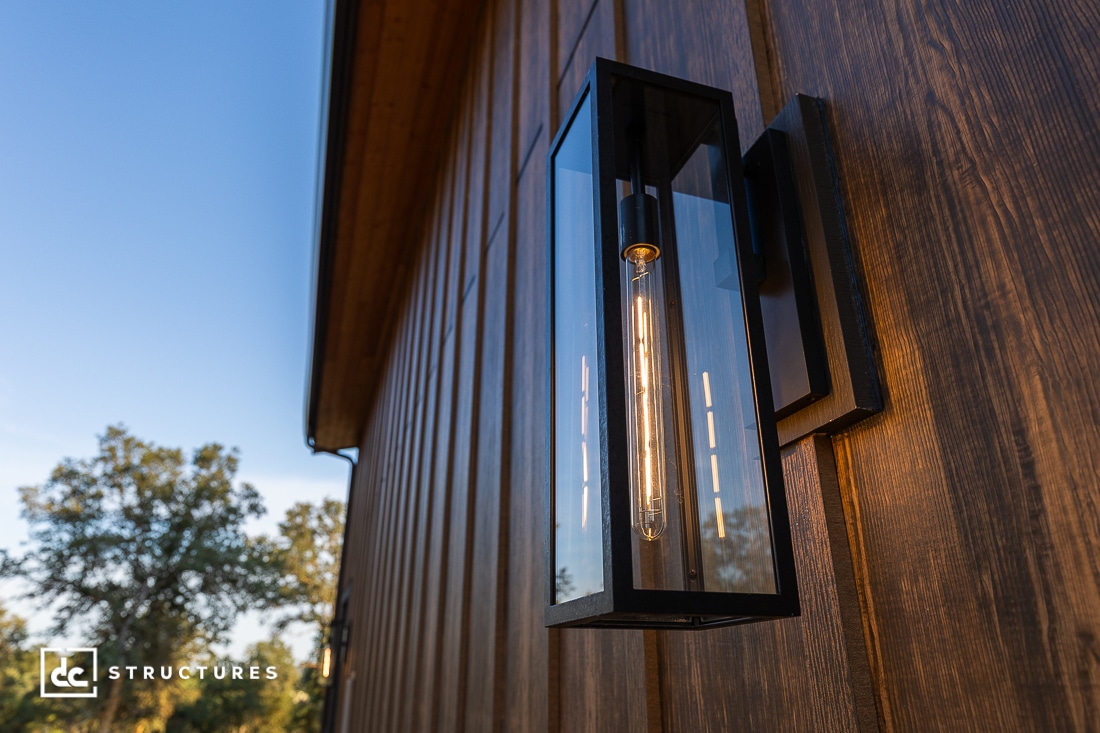 A modern black wall-mounted lantern with a vertical light bulb is attached to a wooden exterior wall in the evening sunlight. Trees and blue sky are visible in the background.
