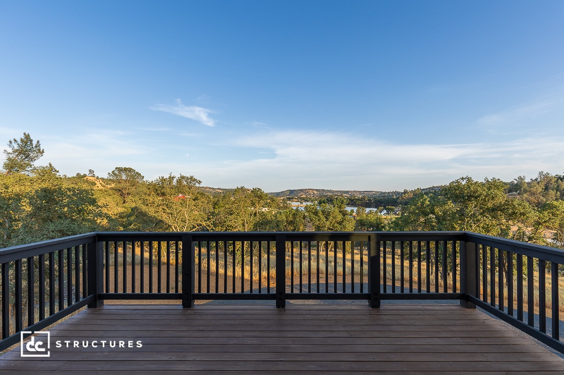 A wooden deck with black railings overlooks a scenic view of green trees, hills, and a clear blue sky under bright daylight.