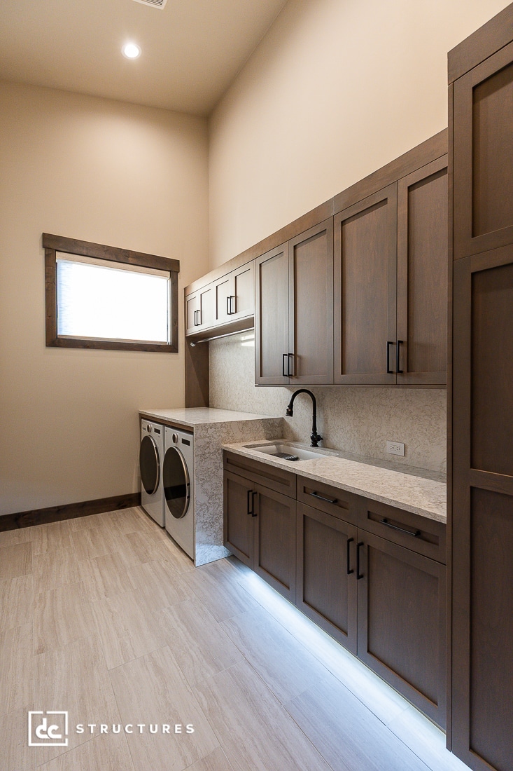 Modern laundry room with wood cabinets, marble countertop, under-cabinet lighting, a front-loading washer and dryer, and a window with natural light. The space has a clean, minimalist design.