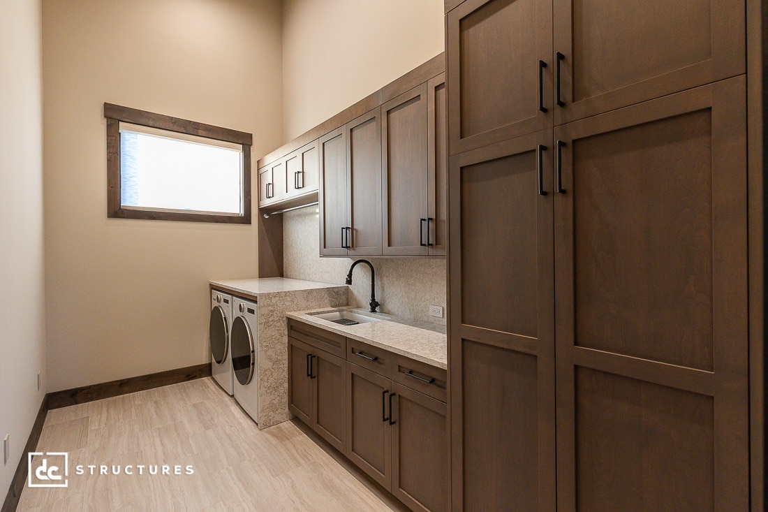 A modern laundry room with wood cabinets, a front-loading washer and dryer, a sink with a black faucet, and a large window letting in natural light. The flooring is light-colored, and the room has a clean, organized look.