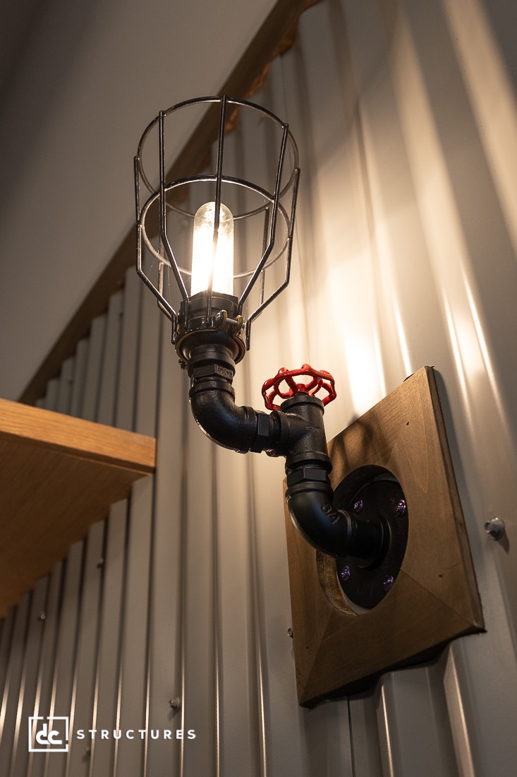 A wall-mounted industrial-style sconce features an exposed bulb with a wire cage, black metal piping, a red valve handle, and a wooden base; it is attached to a corrugated metal wall near a wooden shelf.