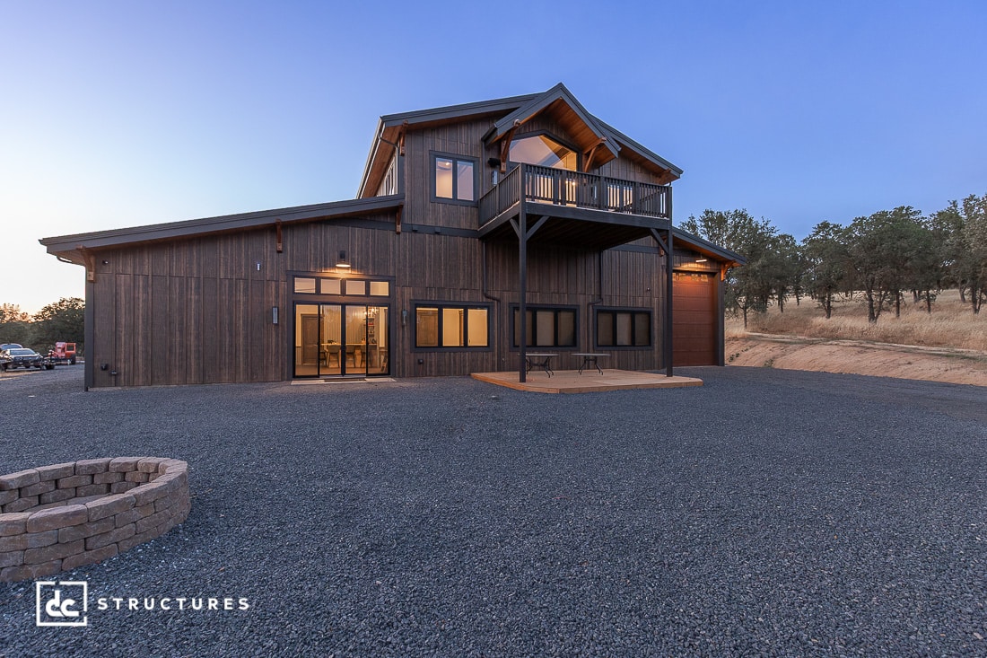 A modern two-story barn-style house with dark wood siding and large windows, set on a gravel lot with a fire pit and surrounded by trees at dusk.