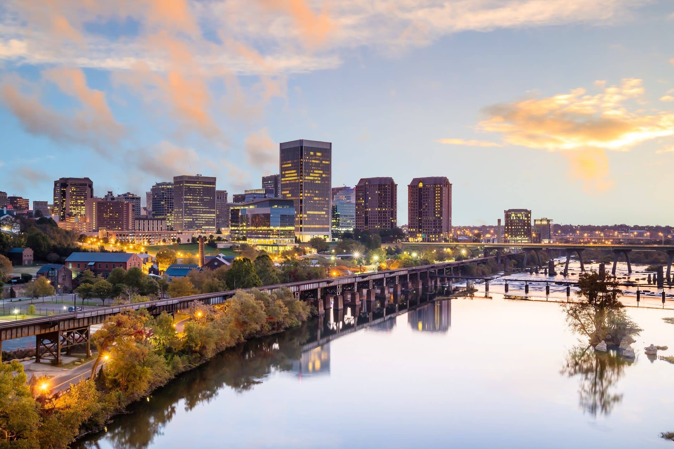 City skyline at dusk with tall modern buildings, a railroad bridge, and river surrounded by trees reflecting the sky with scattered clouds and soft, warm lighting.