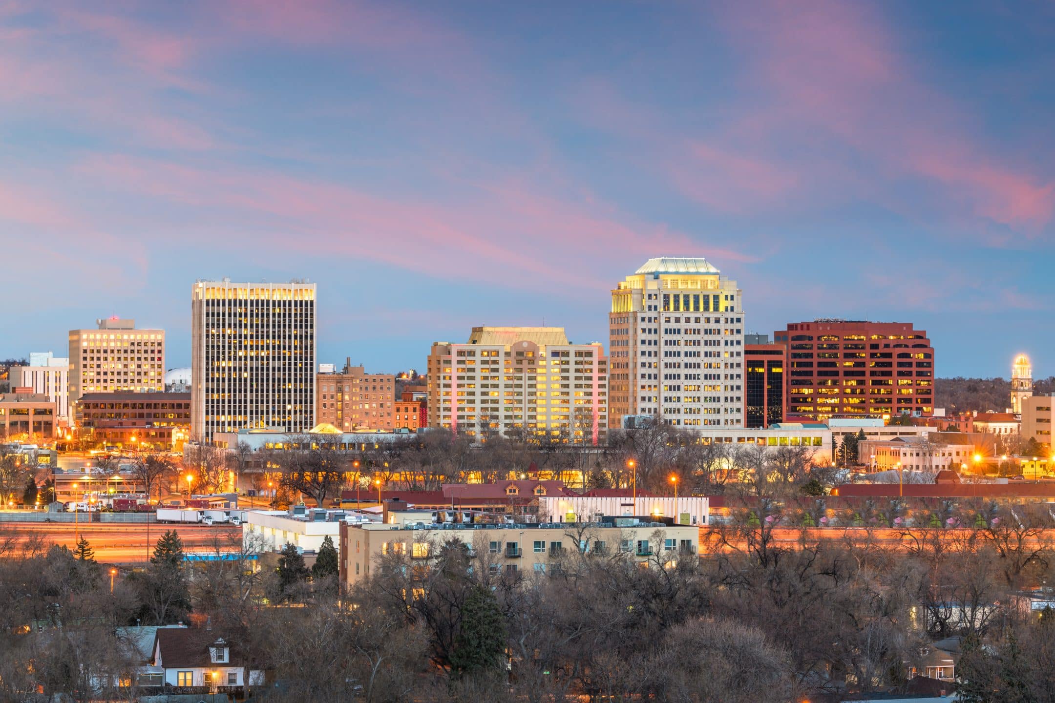 City skyline at sunset with tall buildings and offices illuminated, set against a colorful sky with pink and blue clouds, and trees and smaller houses in the foreground.