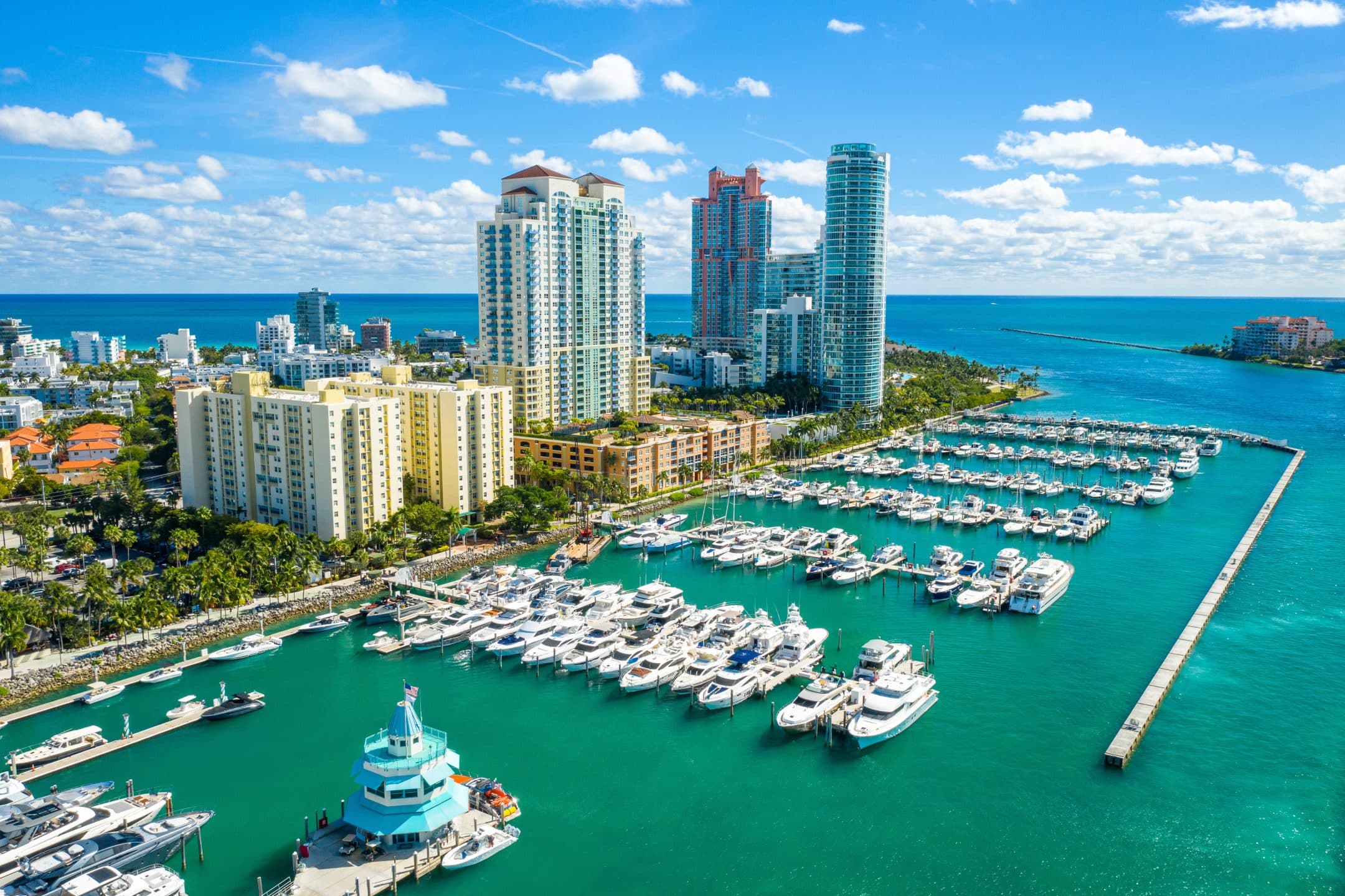 Aerial view of a marina filled with yachts and boats next to tall modern buildings, palm trees, and turquoise water under a bright blue sky in a coastal city.