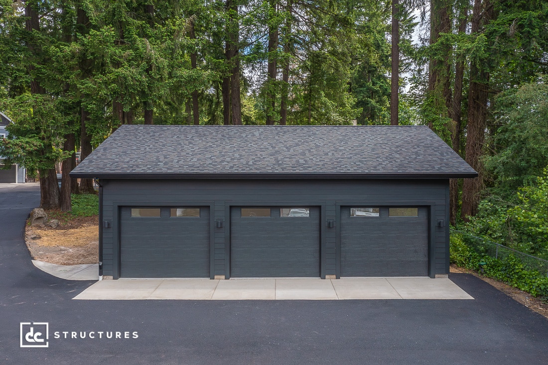 A modern three-car garage with dark gray siding and three matching garage doors sits surrounded by tall trees and greenery. The driveway and sidewalk are clean and neatly paved.