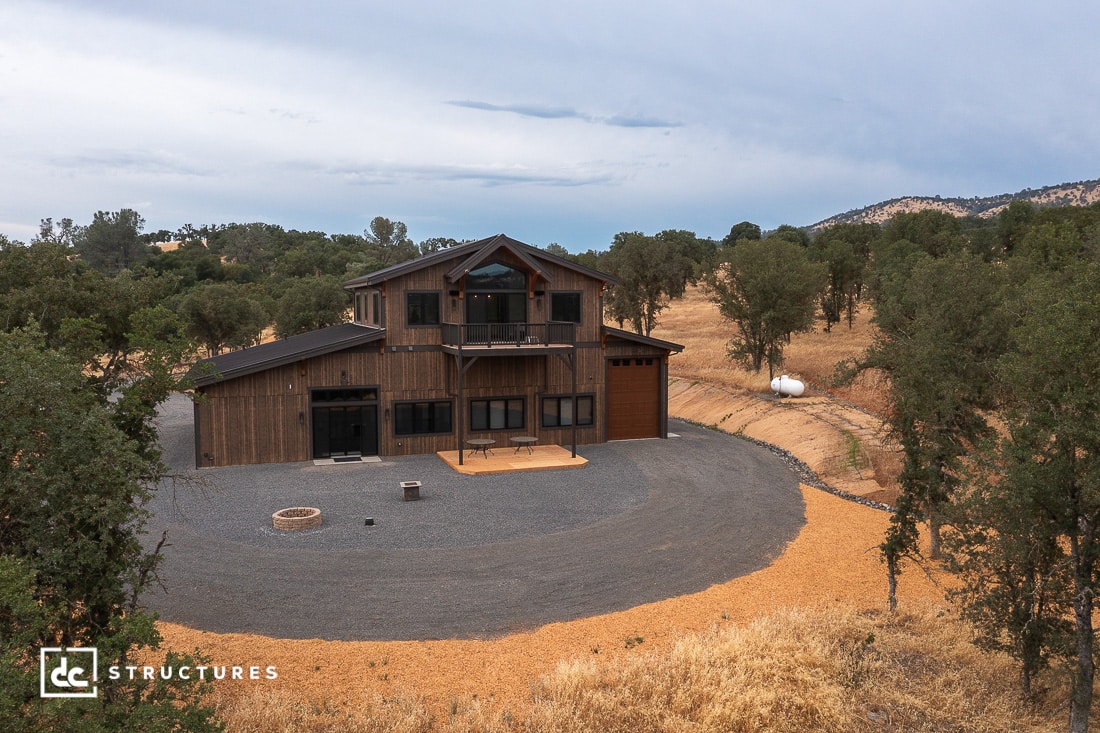 A large, two-story wooden barn with a balcony and garage sits in a rural landscape surrounded by trees and dry grass under a cloudy sky. A gravel driveway circles the front of the building.