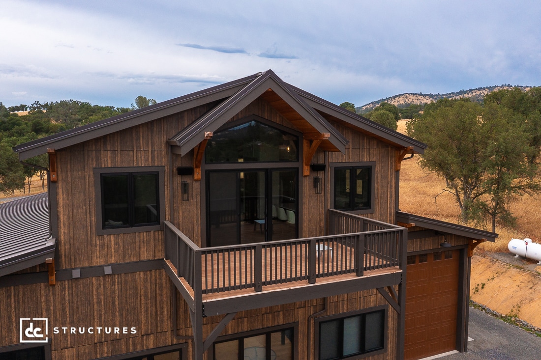 A modern two-story wooden house with a large balcony, black-framed windows, and a scenic background of hills, trees, and dry grass.