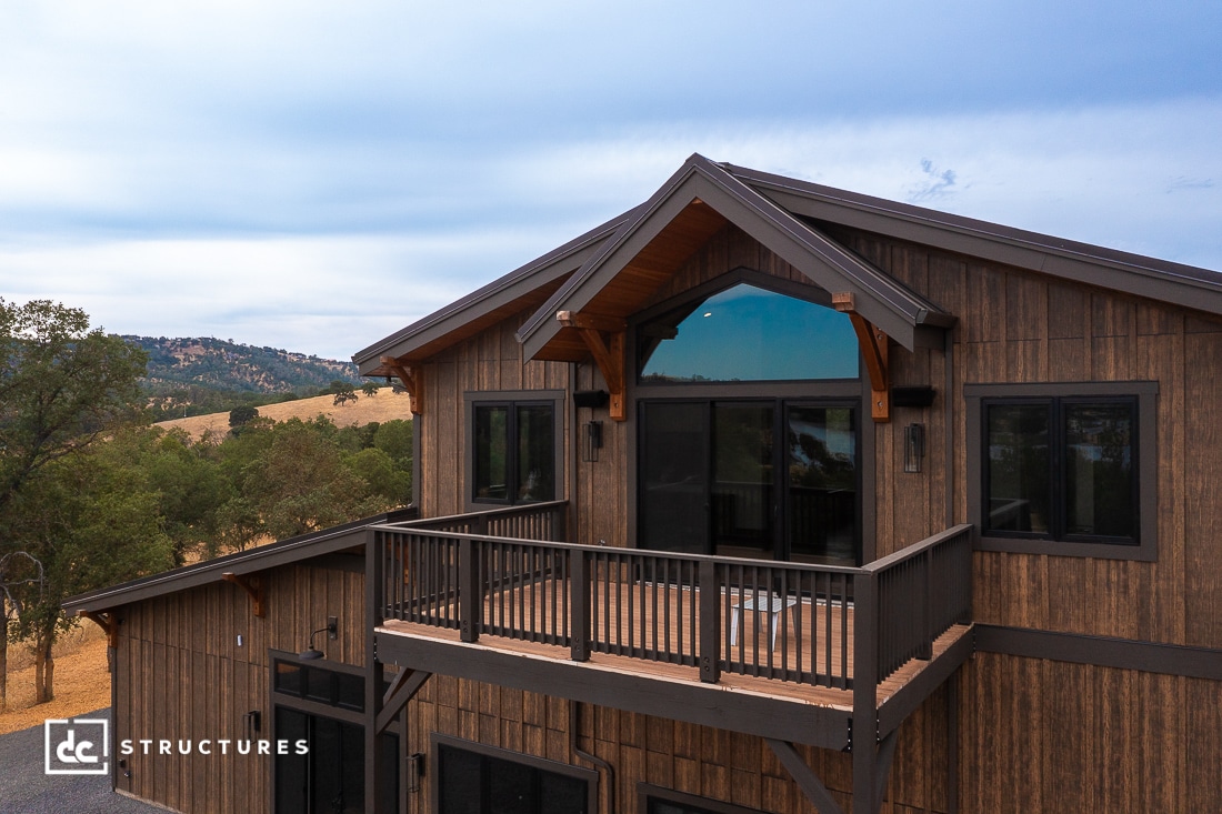 A modern wooden house with large windows and a covered balcony, set in a hilly landscape with trees. The sky is cloudy.