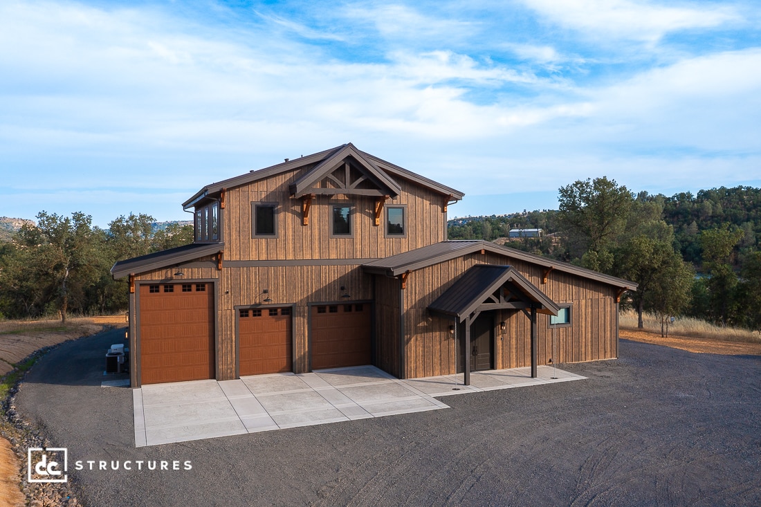 Two-story barn-style building with a wood exterior, brown doors, and a covered entryway, surrounded by trees and hills under a blue sky. Concrete driveway and gravel area in front.
