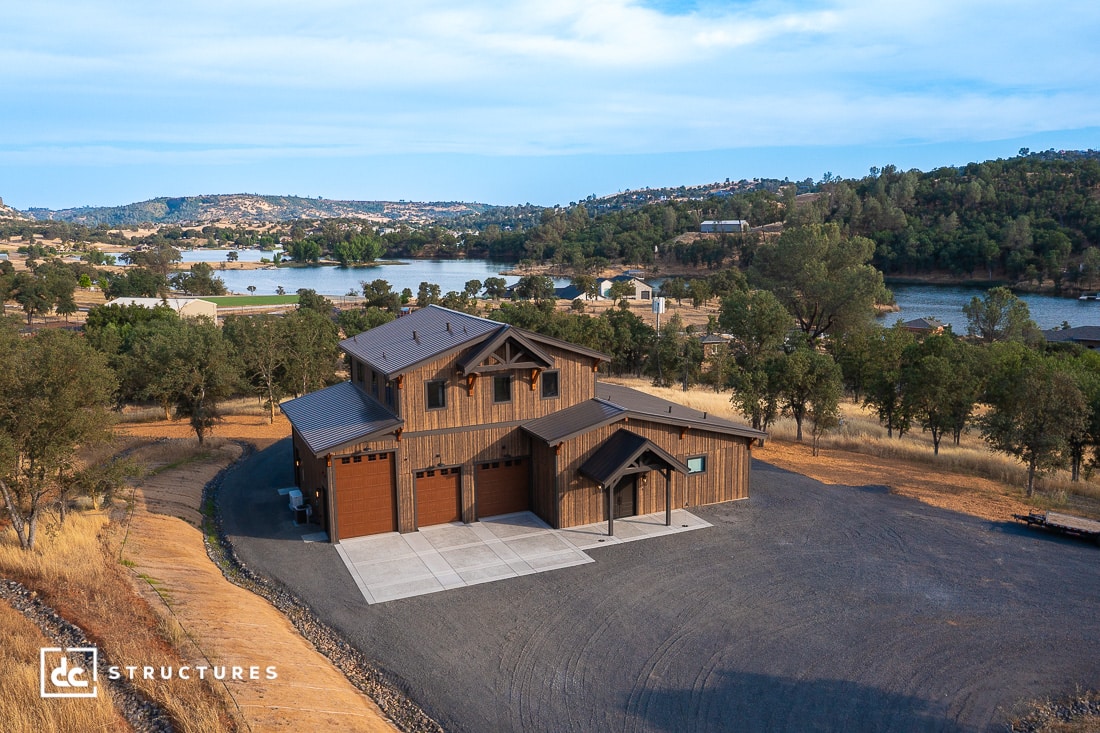 A large wooden house with three garage doors sits on a gravel driveway, surrounded by trees and hills, with a lake and scattered homes visible in the background under a partly cloudy sky.