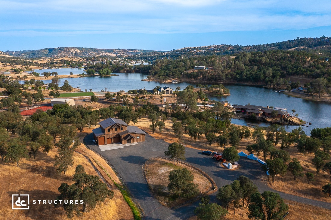 Aerial view of a lakeside property featuring a large wooden house surrounded by trees and dry grass, with several other homes and docks along the shoreline and rolling hills in the background.