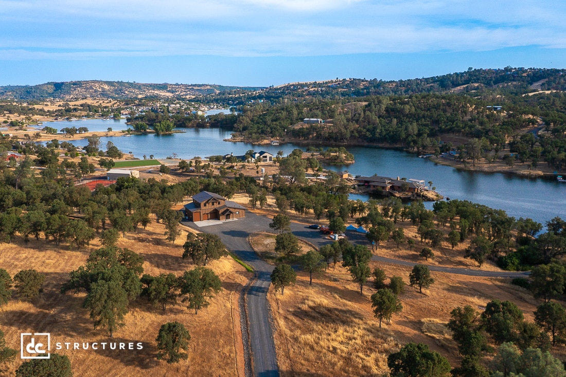 Aerial view of a lakeside rural landscape with scattered houses, trees, and a winding road, surrounded by rolling hills under a partly cloudy sky.