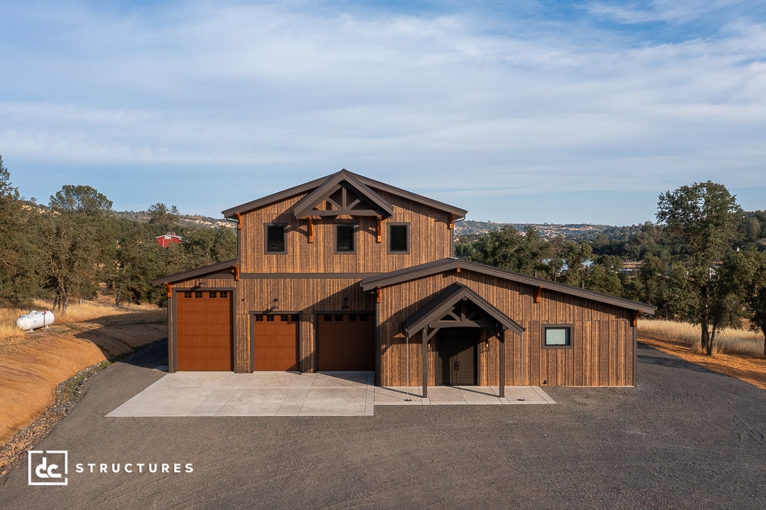 A modern, rustic two-story barn-style building with wood siding, three large garage doors, and a small front entry, set on a concrete pad surrounded by dry grass and trees under a clear sky.