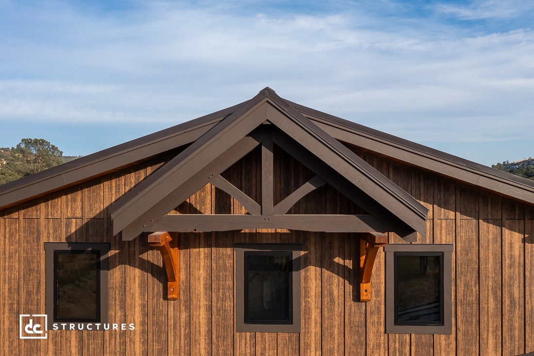 A close-up view of a wooden building’s facade with dark brown trim, three rectangular windows, and exposed timber frame details against a partly cloudy blue sky.