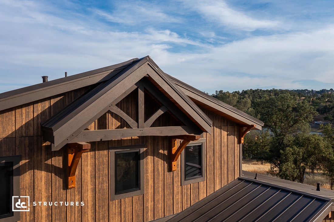 A close-up view of a modern wooden house with a gabled roof, black-framed windows, and decorative timber trusses, set against a backdrop of trees and a blue sky.