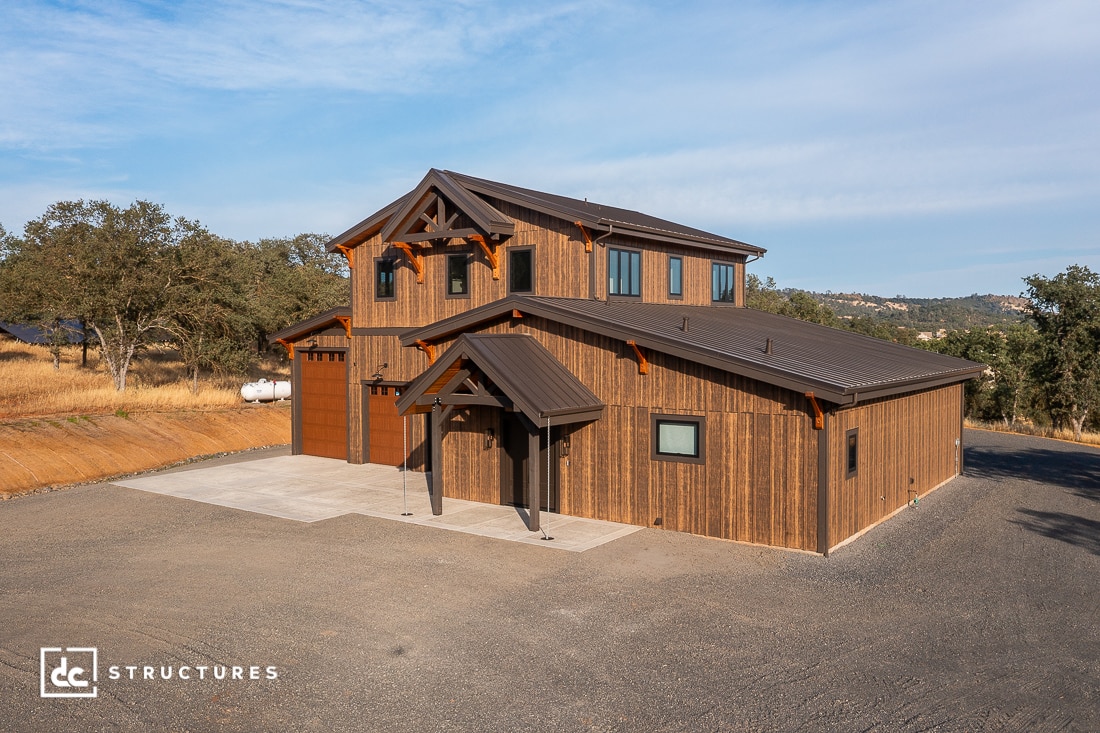 A modern two-story barn-style building with wood siding, a metal roof, and large garage doors sits on a paved lot surrounded by dry grass and trees under a blue sky.