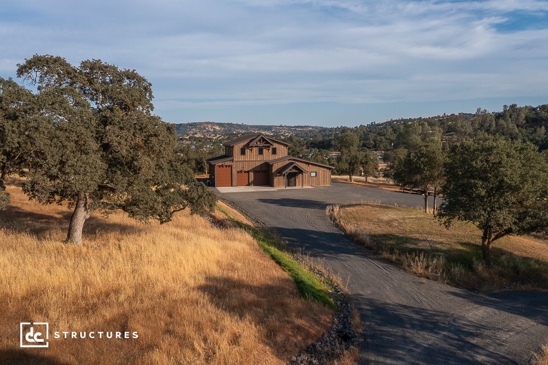 A large wooden house with a three-car garage sits at the end of a paved driveway, surrounded by dry grass, trees, and rolling hills under a blue sky.
