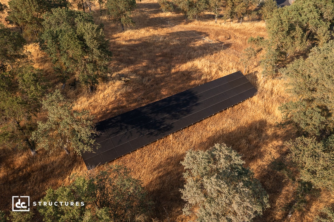 A long, dark-roofed structure sits among tall, dry grass and scattered trees in a rural landscape, viewed from above.