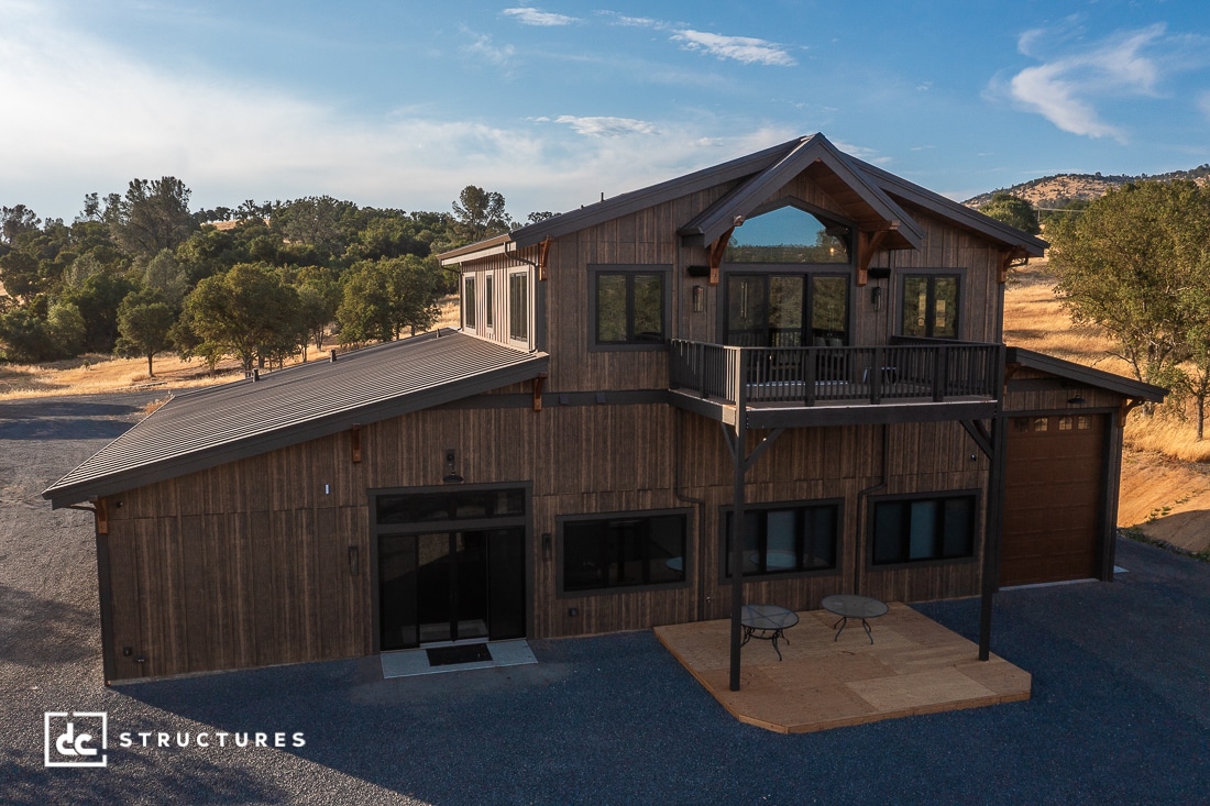 A modern two-story wooden building with large windows, a balcony, and a garage door, surrounded by gravel and dry grassy hills under a blue sky.