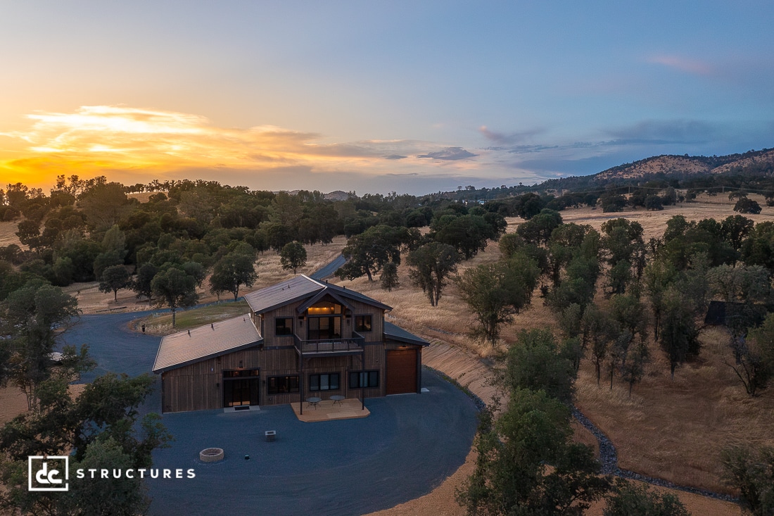 A modern barn-style house sits among trees and dry grasslands at sunset, with hills in the background under a partly cloudy sky.