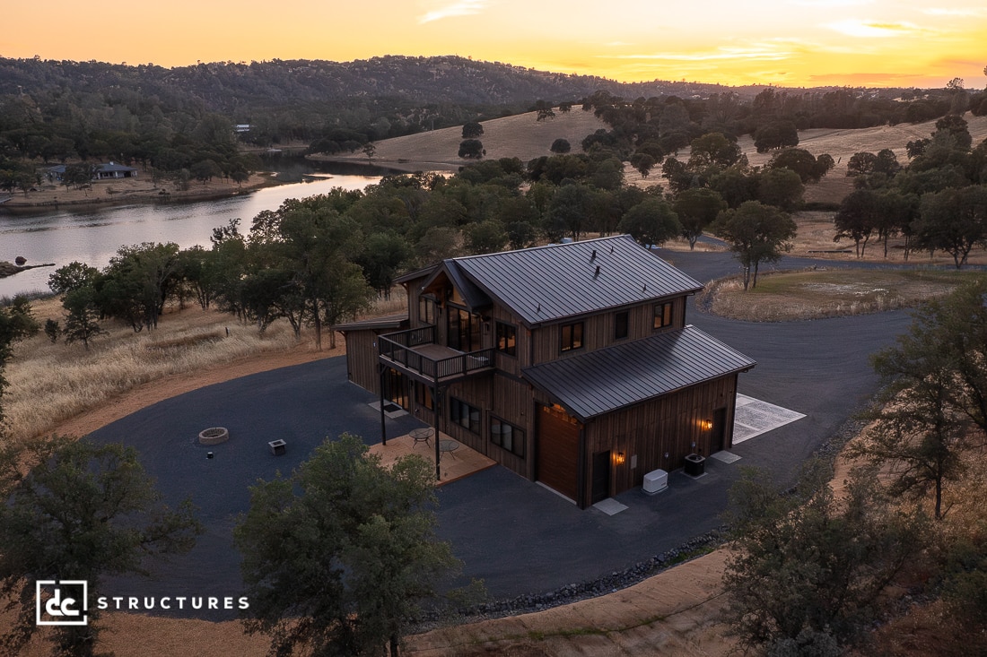 A modern two-story house with dark wood siding and a metal roof sits on a large, circular driveway near a lake, surrounded by rolling hills and trees at sunset.