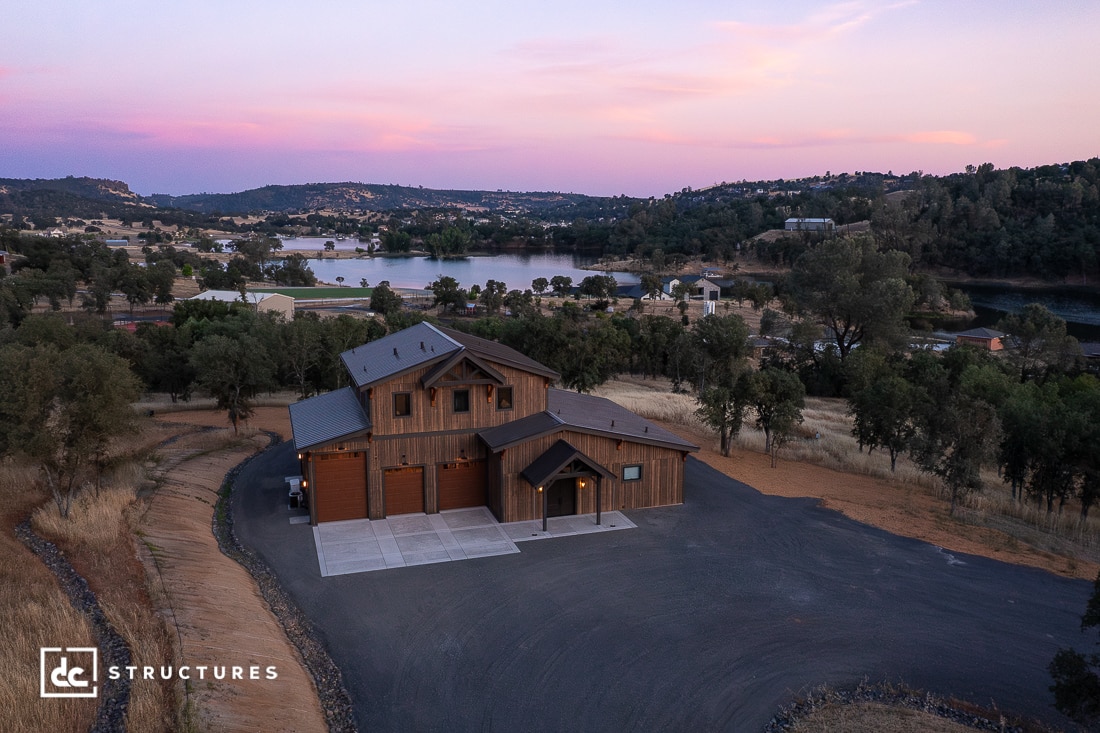 A large wooden house with a multi-car garage sits on a paved driveway, surrounded by trees and rolling hills near a lake at sunset. The sky is tinged with pink and purple hues.