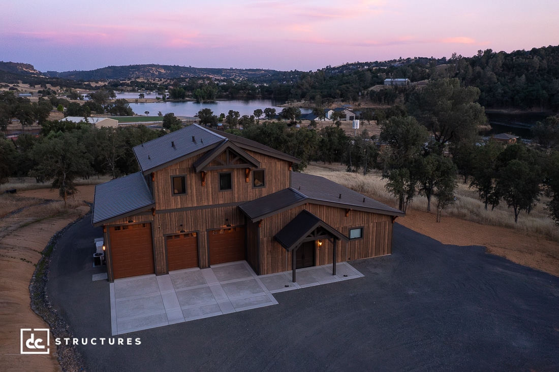 A large modern barn-style house with wood siding and three garage doors sits on a paved lot overlooking a lake and rolling hills at sunset. Trees and distant homes are visible in the background.