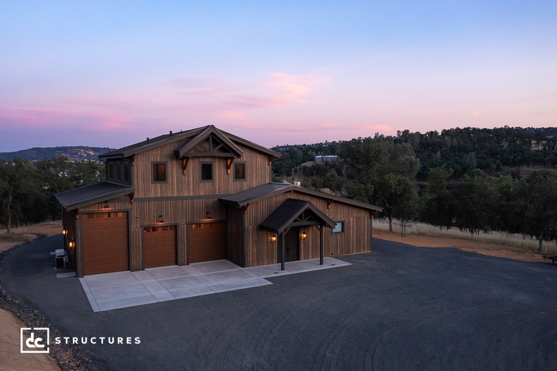 A large, modern barn-style building with wood siding and three garage doors sits on a paved driveway, surrounded by rolling hills and trees at sunset.