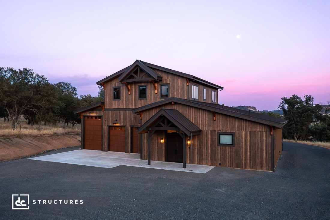 A modern two-story wooden barn with large garage doors, warm exterior lighting, and a sloped roof, set against a twilight sky with trees and distant hills.