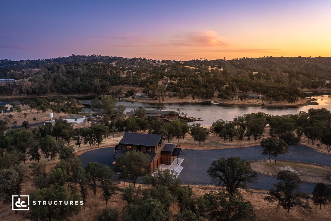 A modern house with a dark roof sits near a winding road by a lake, surrounded by trees and rolling hills at sunset. The sky is tinted with warm hues.