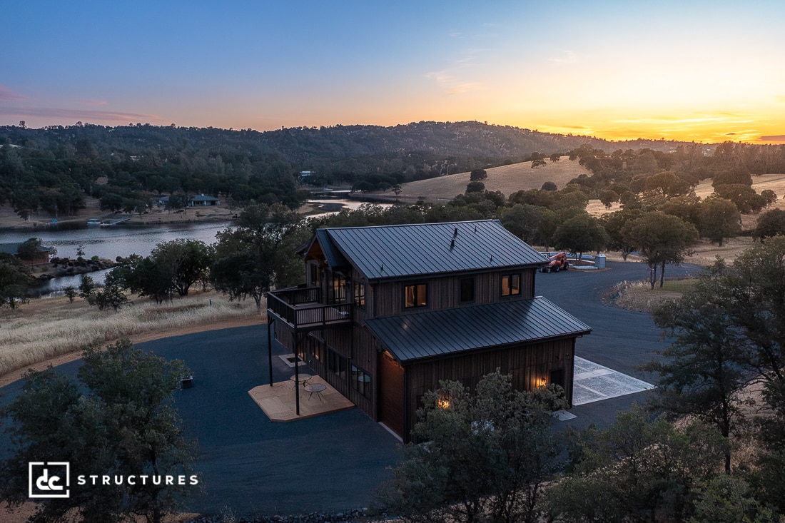 A modern two-story house with a dark metal roof sits surrounded by trees and rolling hills at sunset, near a lake. The sky is colorful, and the house has large windows and a paved driveway.