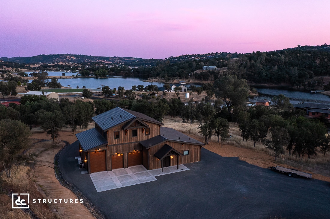 A modern wooden house with exterior lights sits on a paved lot surrounded by trees, overlooking a lake and rolling hills at sunset. The sky is tinged with purple and pink hues.