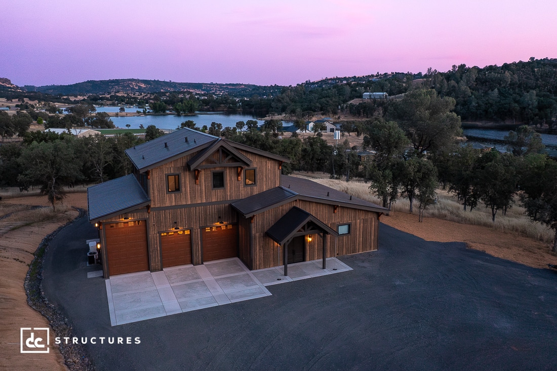 A large, modern wooden barn-style home with three garage doors sits on a paved driveway, surrounded by trees and hills, overlooking a lake at sunset. The sky is purple and pink, creating a peaceful atmosphere.