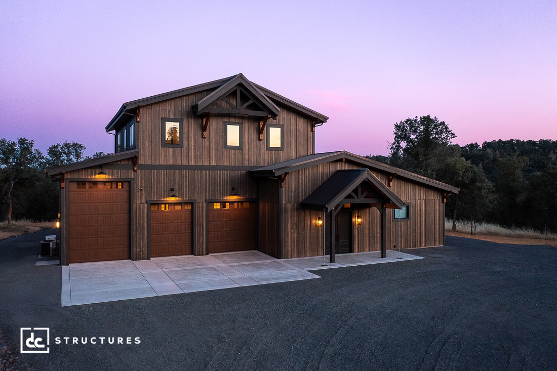 A modern two-story wooden house with three garage doors, warm exterior lights, and a paved driveway, set against a twilight sky with trees in the background. The house features rustic and contemporary design elements.
