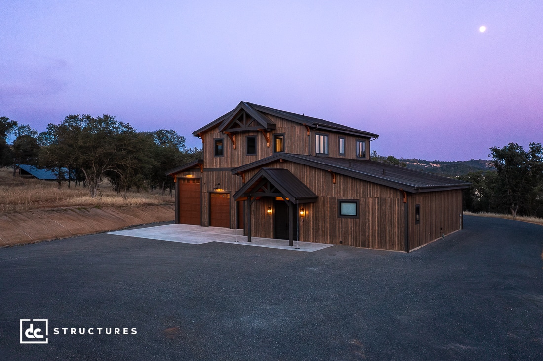 A two-story modern barn-style house with wood siding and large windows, lit warmly at dusk, sits on a spacious lot with a paved driveway and trees in the background.
